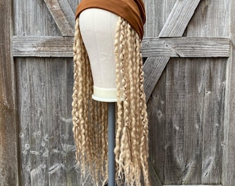 Könnte beinhalten: Ein brauner Turban-Stirnband-Stil mit langen, blonden, lockigen Kunsthaar-Dreadlocks. Die Perücke wird auf einem Schaufensterpuppenkopf präsentiert, der auf einem grauen Ständer montiert ist. Der Hintergrund ist eine verwitterte Holztür.