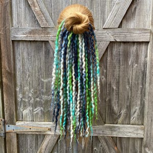 May include: A blonde hair bun with a cascade of colorful dreadlocks. The dreadlocks feature shades of green, blue, and white, creating a vibrant and textured appearance. The hairpiece is displayed against a weathered wooden door.