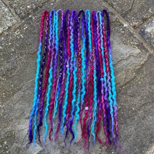 May include: A collection of colorful dreadlocks in shades of blue, purple, pink, and red. Some dreadlocks are adorned with silver wire and small beads. The dreadlocks are arranged in a vertical pattern.