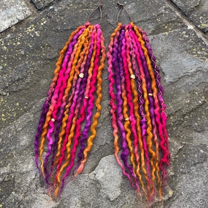 Könnte beinhalten: Zwei handgefertigte Dreadlock-Extensions in leuchtenden Farben. Die Dreads weisen eine Mischung aus Rosa, Orange und Lila auf, mit kleinen goldfarbenen Perlen. Die Dreads sind nebeneinander angeordnet, mit einem braunen Faden oben.