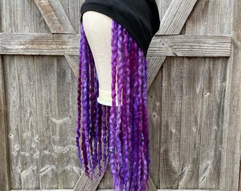 Peut inclure: Un bonnet noir avec de longues dreadlocks violettes et magenta. Les dreadlocks sont coiffées pour pendre, et le chapeau est présenté sur une tête de mannequin. L'arrière-plan est une porte en bois.