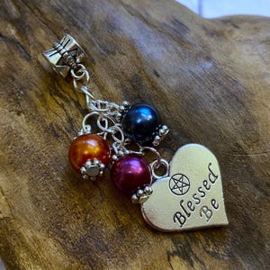 Op de afbeelding: Een zilveren bedel met een hartvormige hanger met de tekst "Blessed Be". De bedel heeft een pentagramsymbool en is verbonden met een ketting met drie kleurrijke kralen: oranje, paars en donkerblauw. De bedel wordt gepresenteerd op een houten oppervlak.