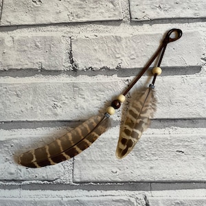 Op de afbeelding: Decoratieve haaraccessoire met twee bruine en tan veren. De veren zijn bevestigd aan een bruin leren koord met houten kralen en zilverkleurige metalen accenten. De veren hebben een natuurlijk, patroonachtig uiterlijk.