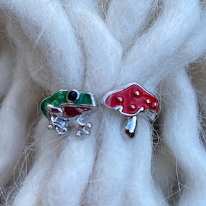 Könnte beinhalten: Zwei kleine Haaraccessoires. Eines ist ein grüner und roter Frosch, das andere ein roter Pilz mit weißem Tupfenmuster. Beide sind aus silberfarbenem Metall. Die Accessoires sind vor einem Hintergrund aus weißen Dreadlocks angeordnet.