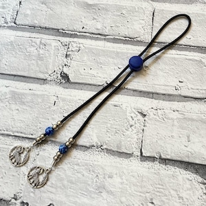 Könnte beinhalten: Ein schwarzes, verstellbares Schlüsselband mit einem blauen Knopf und zwei Strängen. Jeder Strang hat silberne Perlen, blaue und silberne Perlen und einen silbernen Anhänger mit einer Hunde-Silhouette. Das Schlüsselband befindet sich auf einem weißen Ziegelhintergrund.