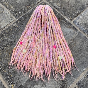May include: A set of synthetic dreadlocks in a blend of pastel pink, lavender, and soft yellow hues. The individual strands feature a twisted texture and are adorned with small gold-toned charms, including stars and leaves, along with pink and green beads.