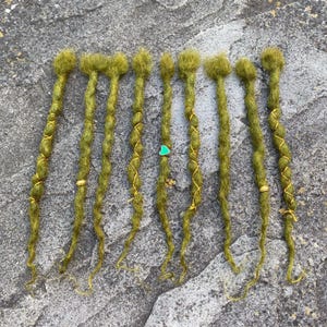 May include: A collection of eight olive green dreadlocks, each with a textured, twisted appearance. Some are adorned with gold-colored wire wraps and small, decorative charms. The dreadlocks are arranged on a gray, stone surface.