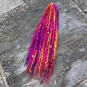Könnte beinhalten: Ein Bündel lebendiger, farbenfroher Dreadlocks in Pink-, Lila- und Orangetönen. Die Dreads sind in einem lockeren, fließenden Stil angeordnet, mit ein paar kleinen, hellfarbigen Perlen dazwischen. Der Hintergrund ist eine strukturierte, graue Steinoberfläche.