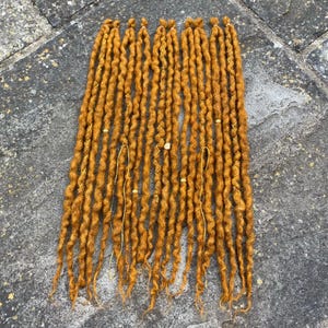 May include: A collection of golden-brown dreadlocks, arranged in a vertical pattern. Each dreadlock is adorned with gold-colored thread and small, decorative beads. The dreadlocks are made of a textured material, likely synthetic or natural hair.