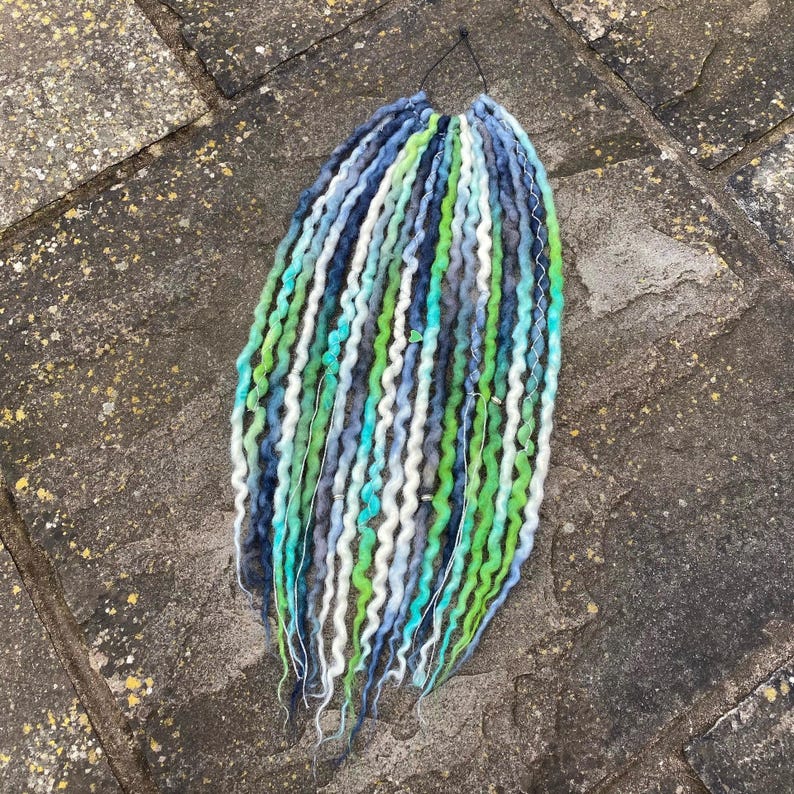 May include: A bundle of colorful dreadlocks in shades of blue, green, and white. The synthetic hair is twisted and textured, with a variety of colors and lengths. The dreadlocks are tied together at the top with a black string.