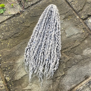 May include: A bundle of gray dreadlocks with black stitching and small blue beads. The dreadlocks are arranged in a vertical pattern, with the ends slightly frayed. The hair accessory is placed on a textured stone surface.