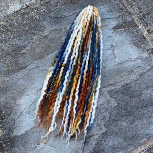 May include: A collection of handmade dreadlocks in shades of white, blue, orange, and yellow. The dreads are decorated with gold thread and wooden beads. The dreadlocks are displayed on a grey stone surface.