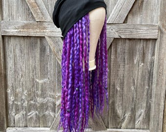 Peut inclure: Un bonnet noir avec de longues dreadlocks violettes et roses. Les dreadlocks sont ornées de petites perles argentées. La perruque est présentée sur une tête de mannequin devant un fond en bois rustique.