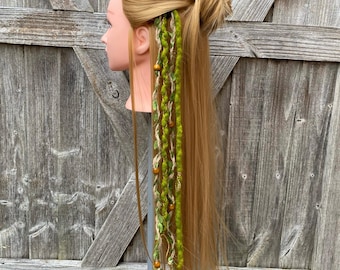May include: A blonde mannequin head with long hair styled in a bun, adorned with green and tan hair wraps. The wraps are decorated with wooden beads and leaf accents, creating a bohemian aesthetic.