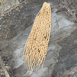 May include: Blonde dreadlocks with wooden beads. The handmade hair extensions are a light, golden colour and are arranged in a vertical pattern. The dreads are lying on a grey, textured surface.