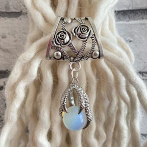 Peut inclure: Une perle de dreadlock argentée avec un motif de rose et une griffe pendante tenant un globe bleu-blanc. La perle est conçue pour être portée avec des dreadlocks ou d'autres coiffures.