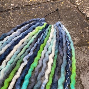 May include: A collection of handmade dreadlocks in various shades of blue, green, and white. The dreads are arranged together and have a silver thread accent. The dreads are made of a soft, textured material.