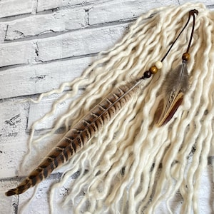 Puede incluir: Dreadlocks de color crema con un cordón de cuero marrón y adornos de plumas. Las plumas son marrones y beige con un patrón natural. Cuentas de madera están unidas al cordón. Las rastas están dispuestas sobre una superficie blanca.
