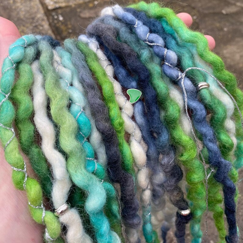 May include: A collection of handmade dreadlocks in shades of green, blue, and white. The dreads are wrapped with silver wire and adorned with small silver beads and a green heart-shaped charm. The overall aesthetic is natural and bohemian.