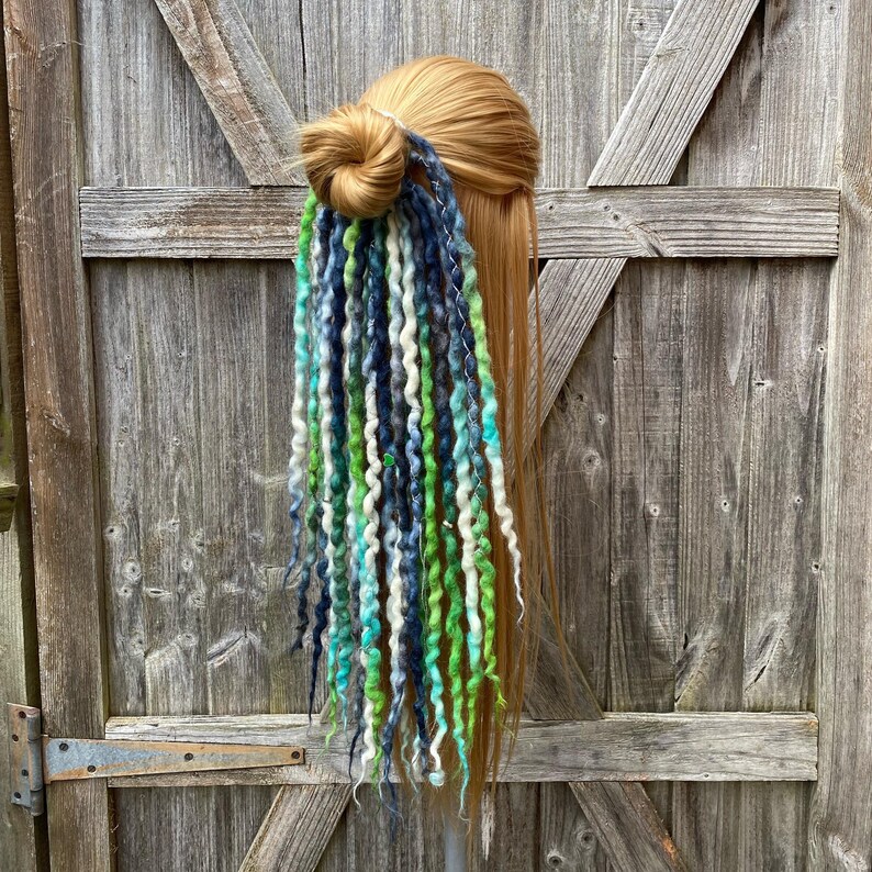 May include: A blonde hair bun with a colorful hair wrap. The wrap features strands of green, blue, and white synthetic dreadlocks. The hair wrap is attached to the bun with a silver wire.