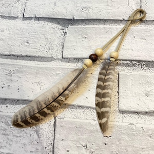 Op de afbeelding: Een decoratief veersieraad met twee veren, houten kralen en een leren koordlus. De veren zijn bruin en wit met een streepjespatroon. De kralen zijn licht- en donkerbruin.