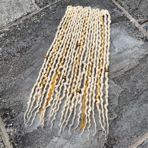 May include: A set of blonde dreadlocks with yellow accents and wooden beads. The dreads are arranged in a neat bundle, showcasing their textured, wavy appearance. The yellow threads and beads add a touch of color and detail to the hair accessory.