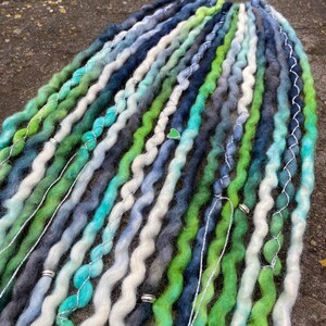 May include: A collection of wavy dreadlocks in shades of green, blue, and white. The dreads are adorned with silver wire and small decorative beads. The color palette includes teal, forest green, and various shades of blue and gray.
