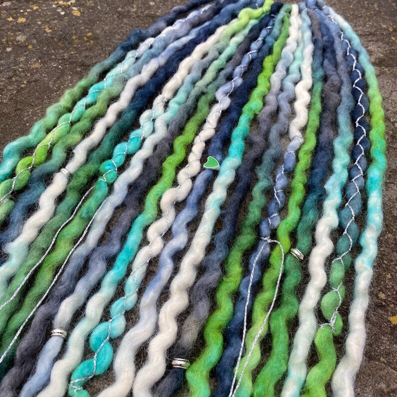 May include: A collection of wavy dreadlocks in shades of green, blue, and white. The dreads are adorned with silver wire and small decorative beads. The color palette includes teal, forest green, and various shades of blue and gray.
