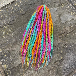 May include: A bundle of colorful dreadlocks in shades of orange, yellow, pink, blue, and green. The dreadlocks are adorned with silver-colored wire and beads. The hair is arranged on a textured, gray stone surface.