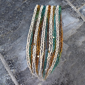 May include: A collection of handmade dreadlocks in various colors, including white, brown, green, and teal. The dreads are adorned with small wooden beads and gold-colored wire accents. The dreadlocks are arranged on a gray stone surface.