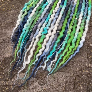 May include: A collection of handmade dreadlocks in various shades of blue, green, and white. The dreads are arranged in a wavy pattern, with some strands featuring silver accents. The overall aesthetic is natural and bohemian.