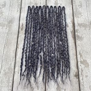 May include: A collection of dark gray dreadlocks, arranged vertically against a weathered wooden background. The dreadlocks are adorned with small silver beads and charms, adding a touch of detail to the textured, handmade appearance. The overall aesthetic suggests a bohemian or alternative style.
