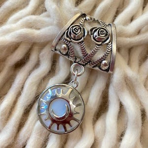 May include: A silver-toned pendant featuring a sun and moon design. The pendant has a circular shape with a sunburst border and a crescent moon in the center. Above the pendant is a decorative piece with two rose designs.
