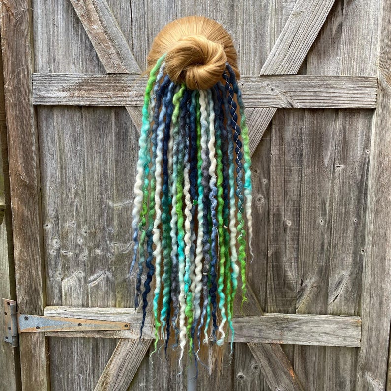 May include: A blonde hair bun with a cascade of colorful dreadlocks. The dreadlocks feature shades of green, blue, and white, creating a vibrant and textured appearance. The hairpiece is displayed against a weathered wooden door.
