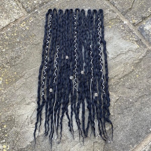 May include: Dark blue dreadlocks with white accents and decorative silver beads. The dreadlocks are arranged in a rectangular pattern, showcasing their texture and style. The image is taken from a top-down perspective, highlighting the intricate details of the hair extensions.