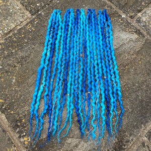May include: A collection of vibrant blue dreadlocks with varying shades of turquoise and light blue. Each dreadlock is adorned with silver wire and small silver beads. The dreadlocks are arranged on a textured, gray surface.