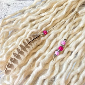 Puede incluir: Dos accesorios para el cabello de estilo bohemio. Uno presenta una pluma marrón y blanca con cuentas rosas. El otro tiene cuentas rosas y plateadas. Ambos están unidos a un cordón de cuero beige. El fondo es una masa de rastas color crema.