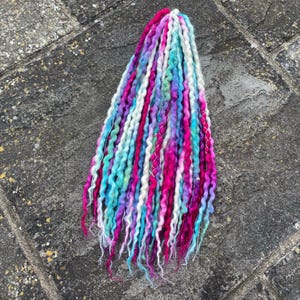 Può includere: Un fascio di dreadlocks colorati nelle tonalità del rosa, viola, blu, bianco e verde. I capelli sintetici sono attorcigliati e acconciati, con un mix di tonalità vivaci e pastello. I dreadlocks sono disposti in un fascio ordinato.