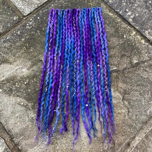 May include: A collection of handmade dreadlocks in shades of blue and purple. The dreads have a wavy texture and are adorned with silver wire and small beads. The color gradient creates a vibrant and unique look for hair styling.