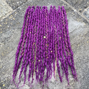 May include: A collection of vibrant purple dreadlocks, arranged on a textured surface. Each dreadlock is adorned with gold-colored accents, including beads and wrapped thread, adding a touch of elegance to the handmade hair extensions.