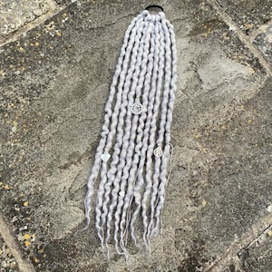 May include: A set of light gray synthetic hair extensions attached to a black elastic hair tie. The long, twisted strands feature silver-toned metal charms, including a five-pointed star, a heart shape, and a circular filigree design.