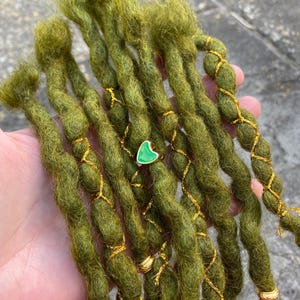 May include: Close-up of olive green dreadlocks, adorned with gold-colored wire and small gold beads. A green heart-shaped charm is visible. The dreadlocks are thick and textured, suggesting a handmade or natural style.