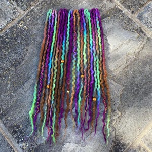 May include: A collection of colorful dreadlocks in shades of purple, green, blue, and brown. The dreads are adorned with gold wire and small beads. The dreadlocks are arranged on a gray stone surface.