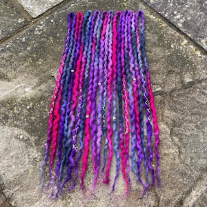 May include: A collection of vibrant, handmade dreadlocks in shades of purple, pink, and blue. The dreads are adorned with silver accents and are arranged against a textured background. These are designed for hair styling.