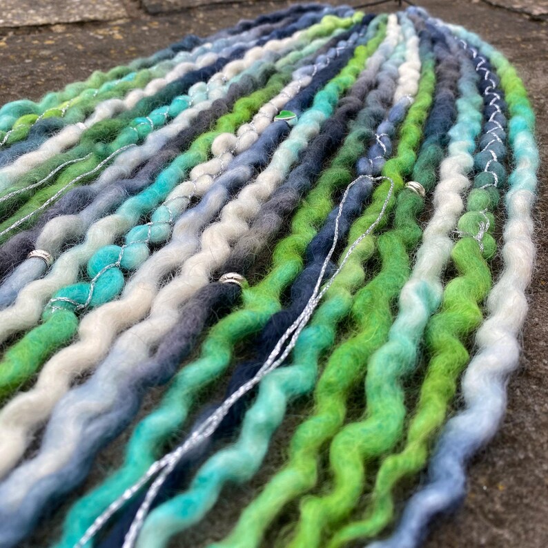 May include: A collection of handmade dreadlocks in shades of green, blue, and gray. The dreads are adorned with silver wire and small metal beads. The color palette creates a natural, ocean-inspired aesthetic.