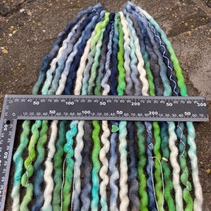 May include: A collection of handmade dreadlocks in shades of blue, green, white, and gray. The dreads are arranged in a bundle, with a ruler placed across them for measurement. The dreadlocks appear to be made of wool or a similar fiber.