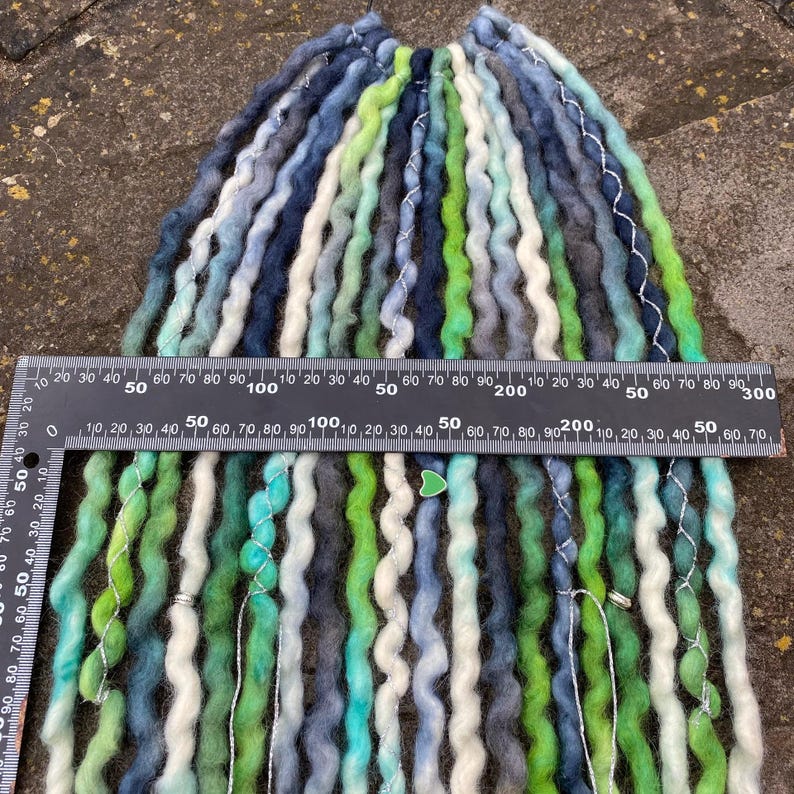 May include: A collection of handmade dreadlocks in shades of blue, green, white, and gray. The dreads are arranged in a bundle, with a ruler placed across them for measurement. The dreadlocks appear to be made of wool or a similar fiber.