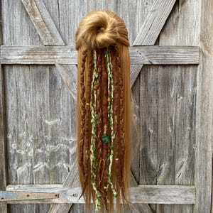Puede incluir: Una cabeza de maniquí con cabello largo castaño claro peinado en un moño, adornada con rastas marrones y crema. Las rastas están decoradas con hojas verdes, cuentas de madera y detalles dorados, creando una estética bohemia.