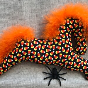May include: A black and orange plush horse with a candy corn pattern and orange feather mane and tail. A black spider is in the foreground.