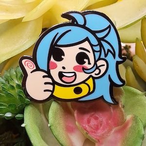 May include: A colorful enamel pin featuring a cartoon character with light blue hair and a yellow collar giving a thumbs-up gesture. The pin has black outlines and pink blush on the cheeks. The background is a mix of green and pink plants.